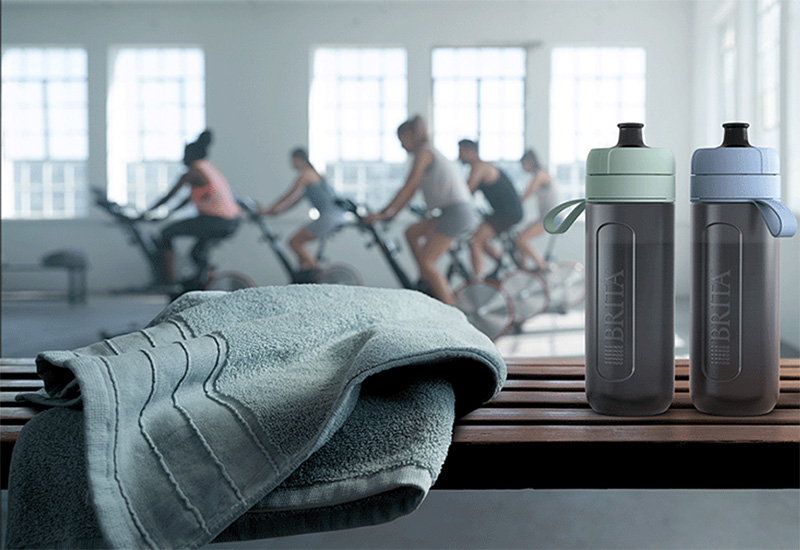 Model Active in blau und grün Zwei Model Actives stehen auf einer Bank im Fitnessstudio, dahinter findet ein Spinning-Kurs statt.