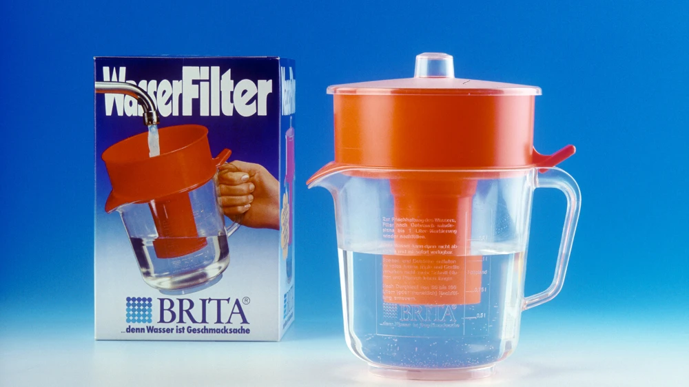 Historischer BRITA Tischwasserfilter von 1970 mit Originalverpackung vor blauem Hintergrund.