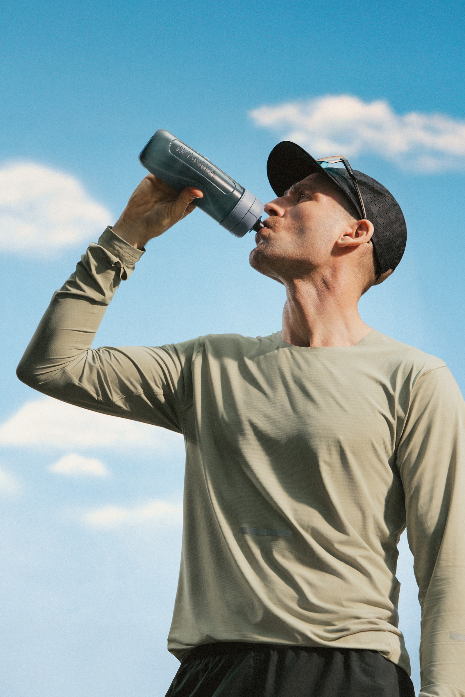 Sportlicher Läufer trinkt aus einer Trinkflasche unter freiem Himmel vor blauem Himmel mit Wolken.
