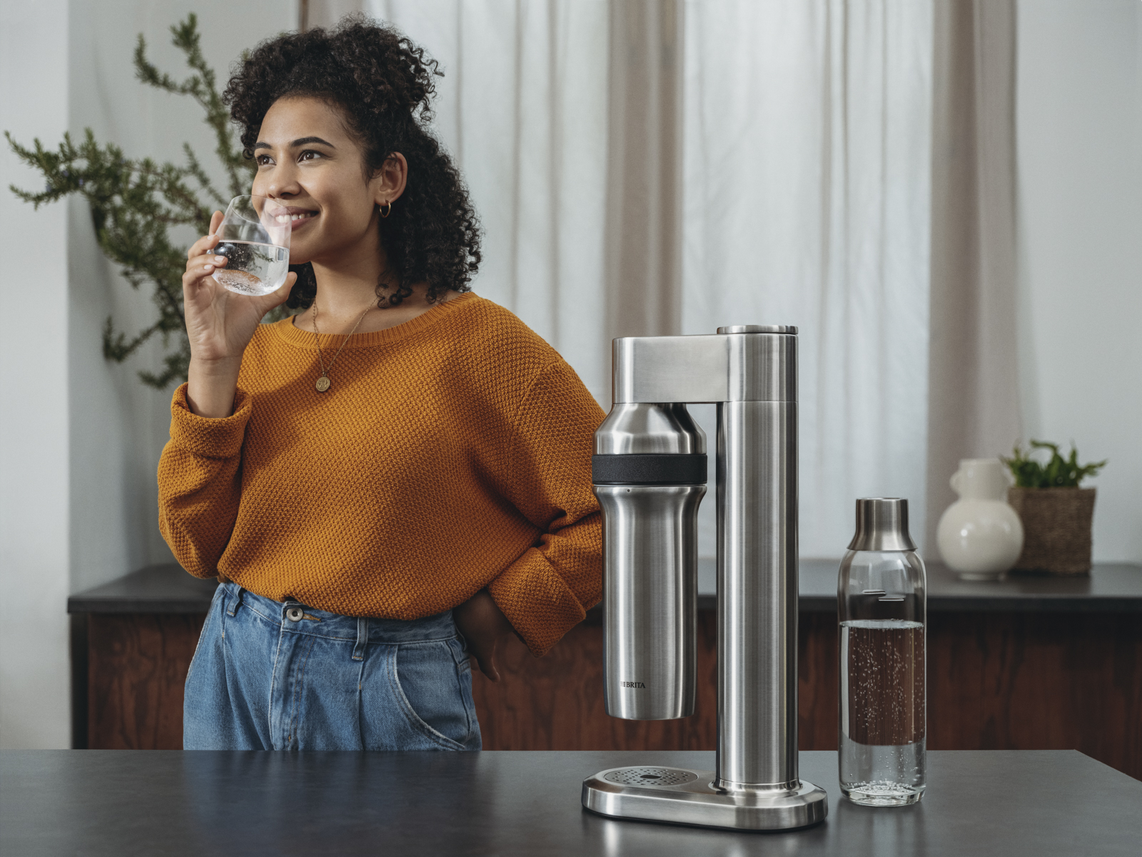Frau trinkt ein Glas Wasser neben sodaTRIO pro. Eine Frau trinkt ein Glas Wasser neben dem sodaTRIO pro Wassersprudler.