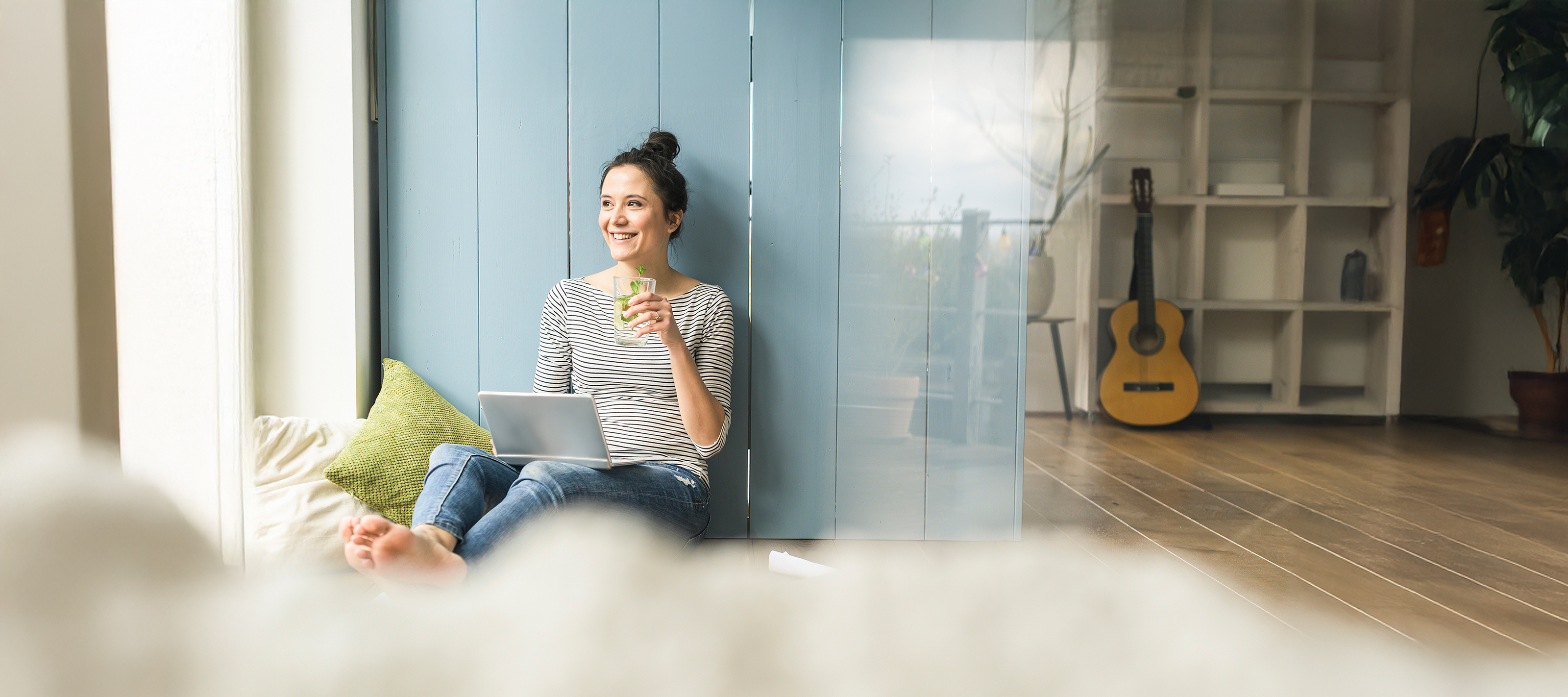 Frau mit Laptop lächelt mit einem Glas Wasser in der Hand Eine Frau sitzt auf Kissen, lächelt und hält ein Glas Wasser in ihrer Hand