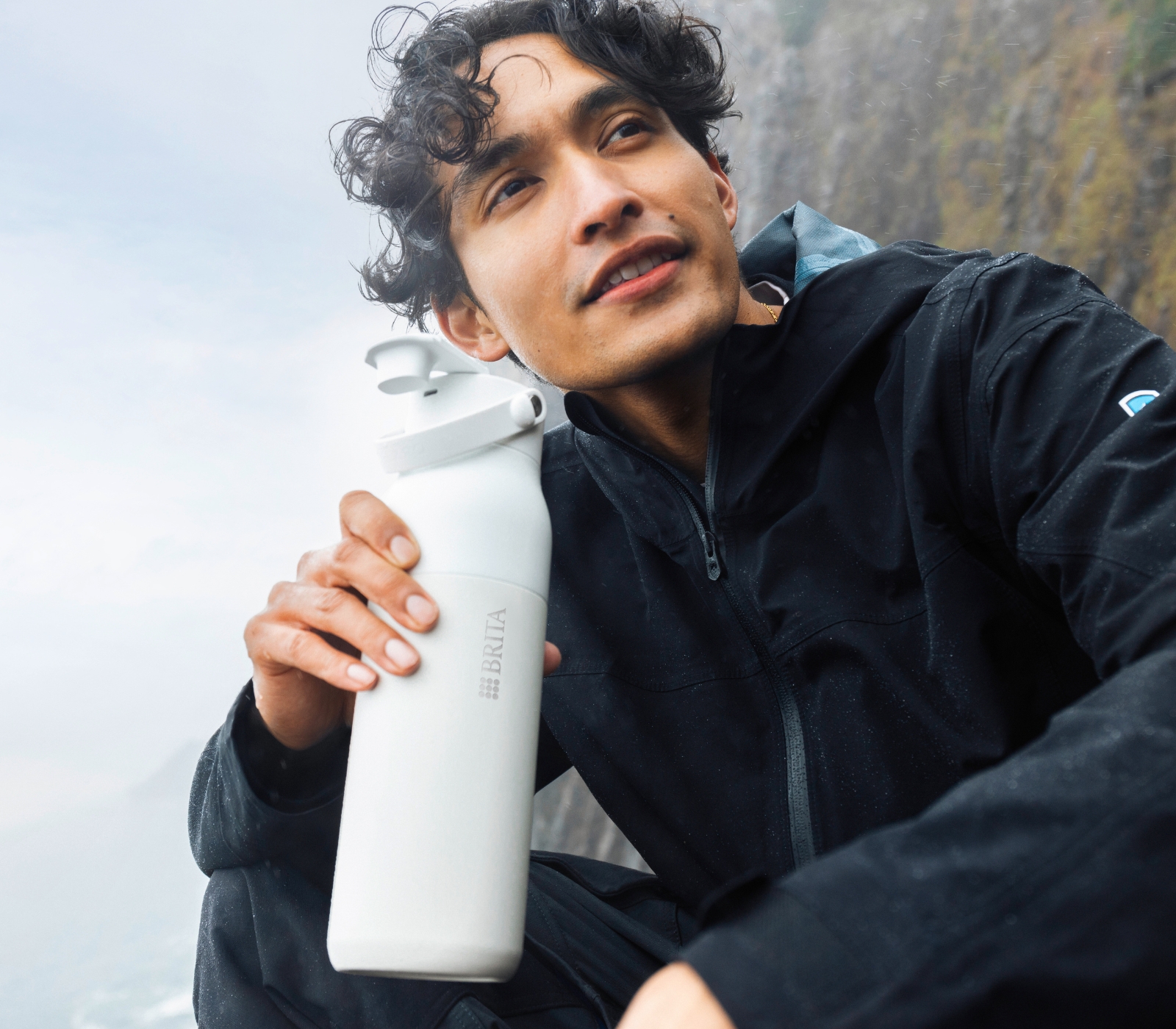 Ein junger Mann in schwarzer Kleidung sitzt in der Natur und trinkt aus der BRITA LARQ iQ Flasche.
