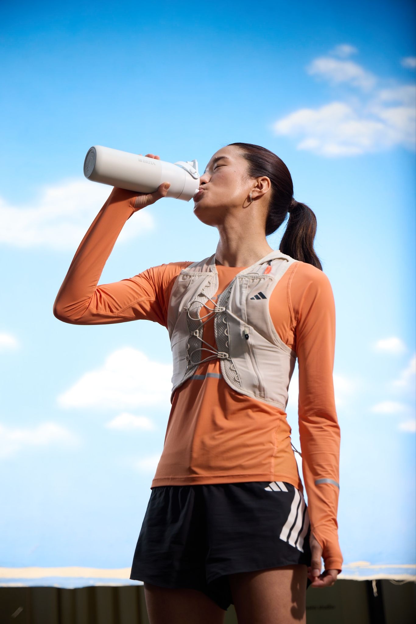 Sportlerin trinkt aus einer weißen BRITA‑Trinkflasche Sportlerin trinkt aus einer weißen BRITA‑Trinkflasche im Freien vor blauem Himmel