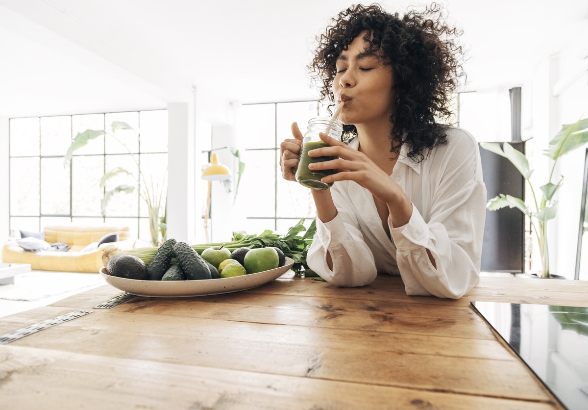 Detox Kur: Detox und Fasten für jeden Lebensstil Frau mit braunen Locken sützt sich auf Holztisch und trinkt einen grünen Smoothie aus einem Glas