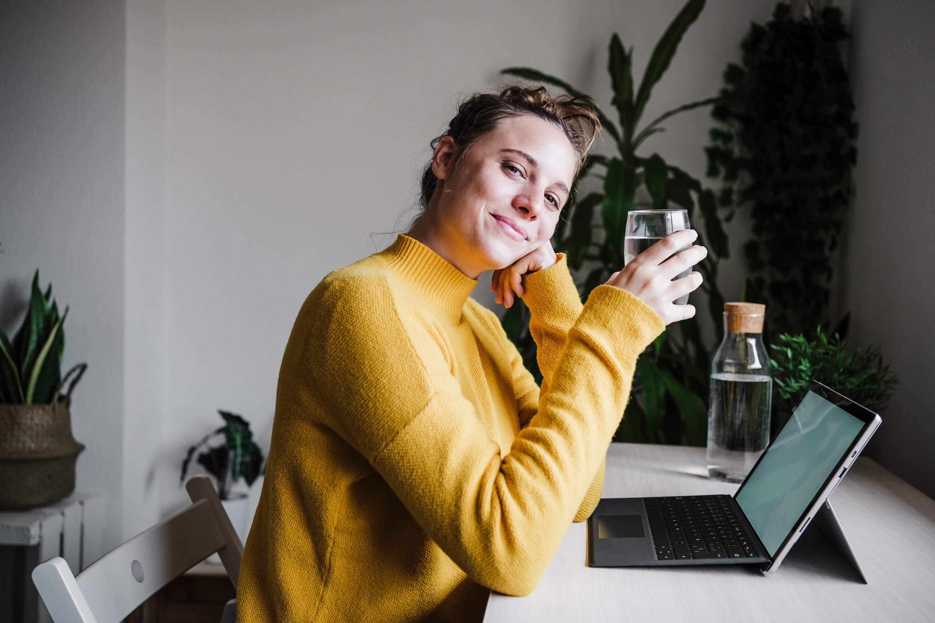 Frau im gelben Pulli trinkt Wasser am Schreibtisch Eine junge Frau mit gelben Pulli sitzt am Schreibtisch und trinkt ein Glas Wasser