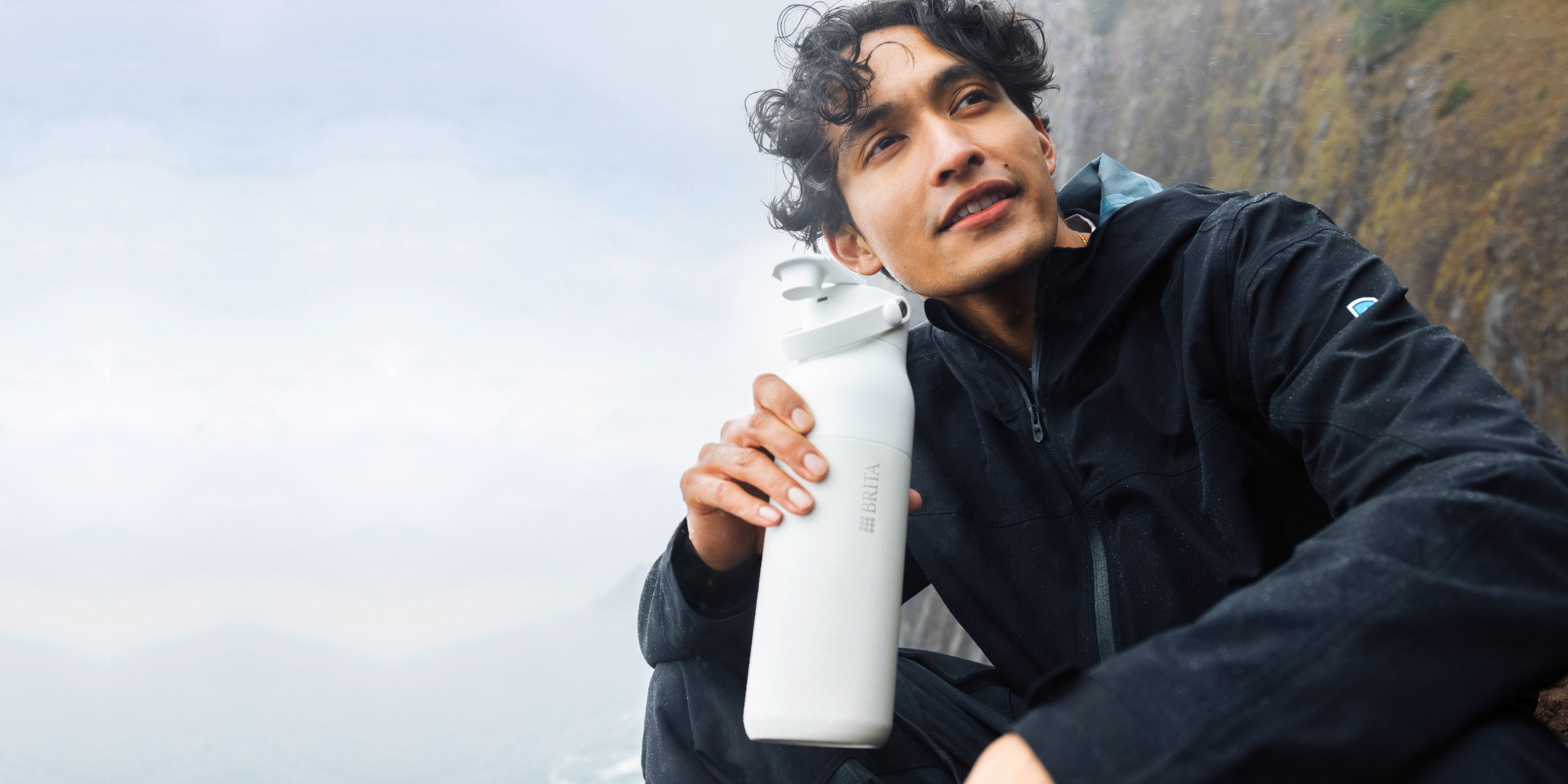 Die BRITA LARQ iQ immer dabei Ein junger Mann in schwarzer Kleidung sitzt in der Natur und trinkt aus der BRITA LARQ iQ Flasche.