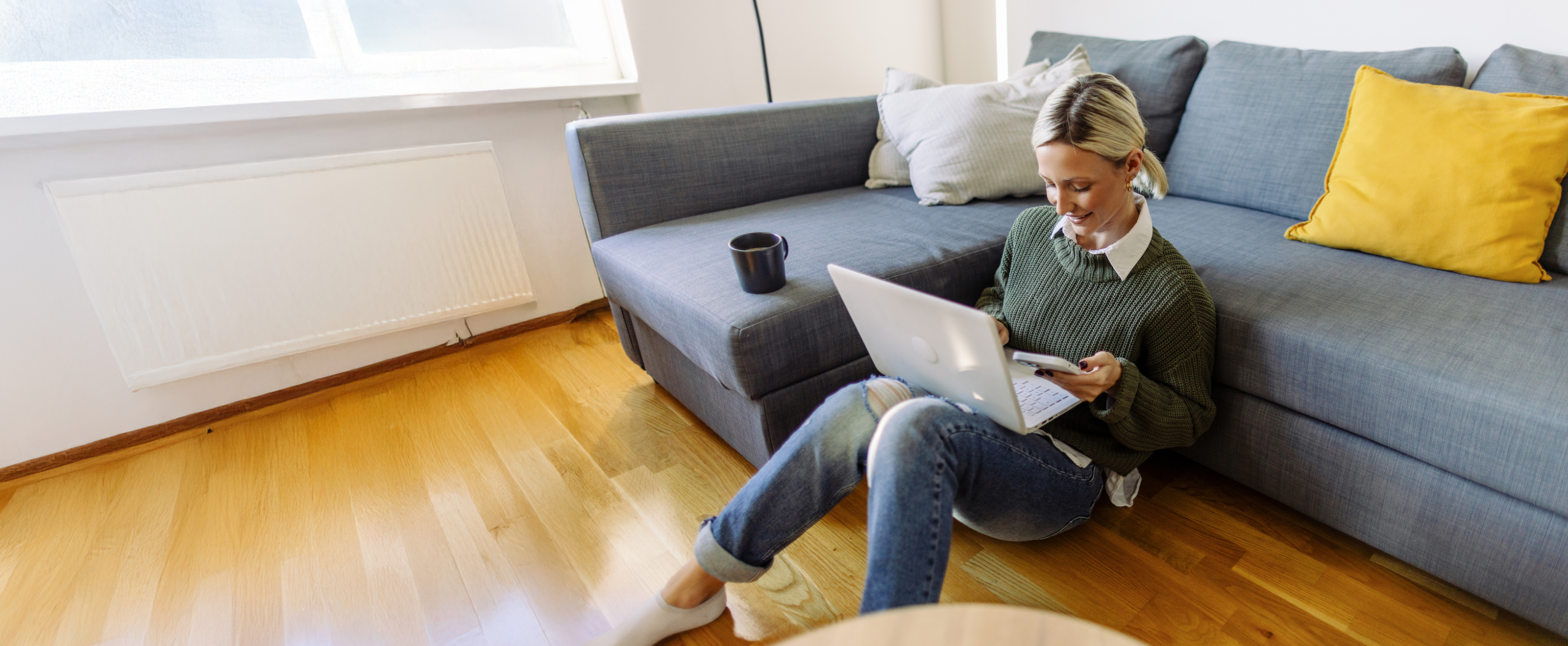 Frau sitzt lächelnd mit Laptop und Smartphone auf Boden Eine junge Frau sitzt lächelnd mit ihrem Laptop und Smartphone auf dem Boden vor der Couch.
