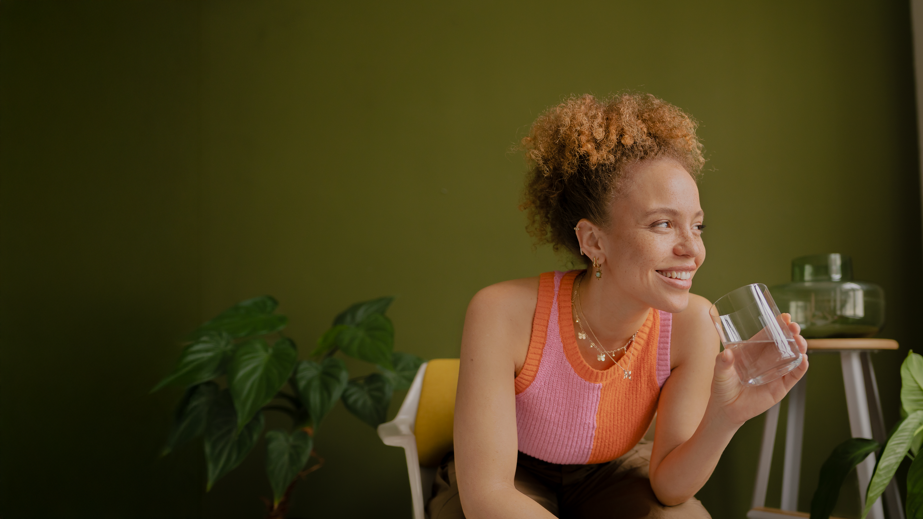 Eine Frau sitzt lächelnd auf einem Stuhl mit einem Glas Wasser in der Hand