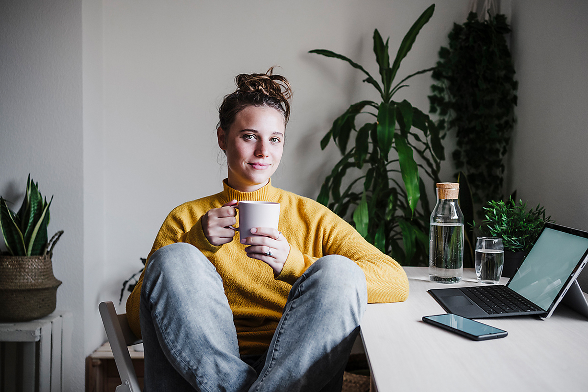 Eine junge Frau sitzt gemütlich an ihrem Schreibtisch und trinkt Kaffee
