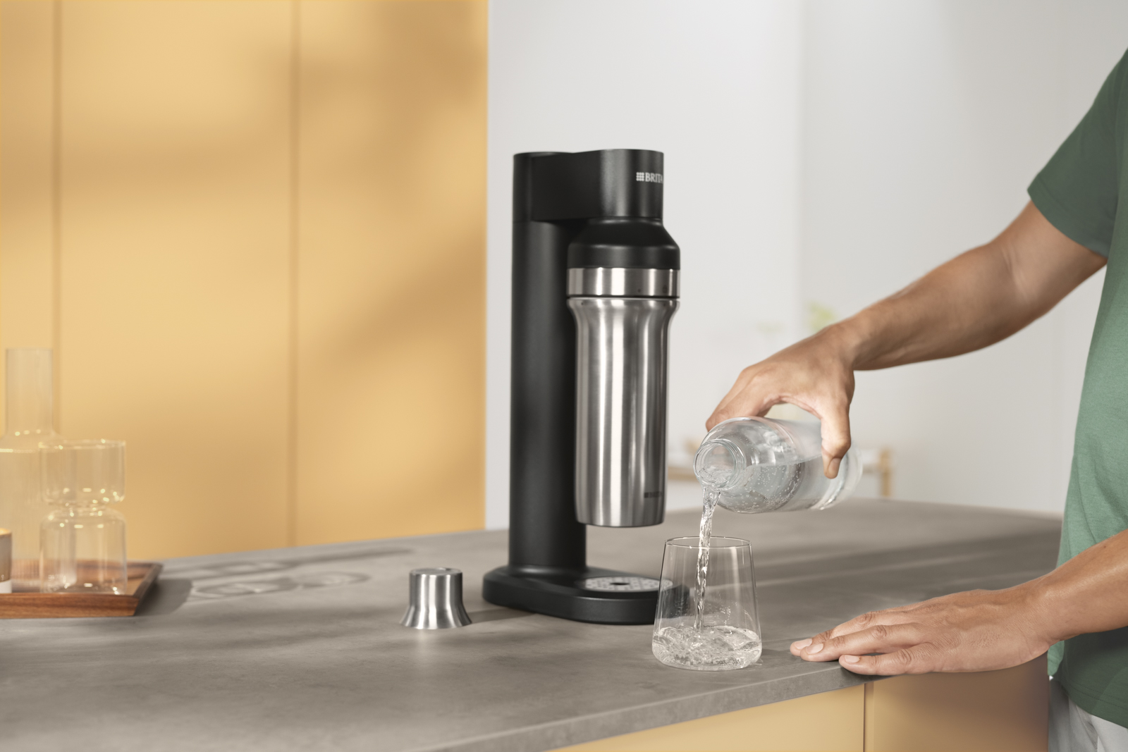 Mann gießt ein Glas Wasser neben BRITA sodaTRIO ein Ein Mann gießt sich ein Glas Wasser ein und steht neben dem BRITA sodaTRIO.