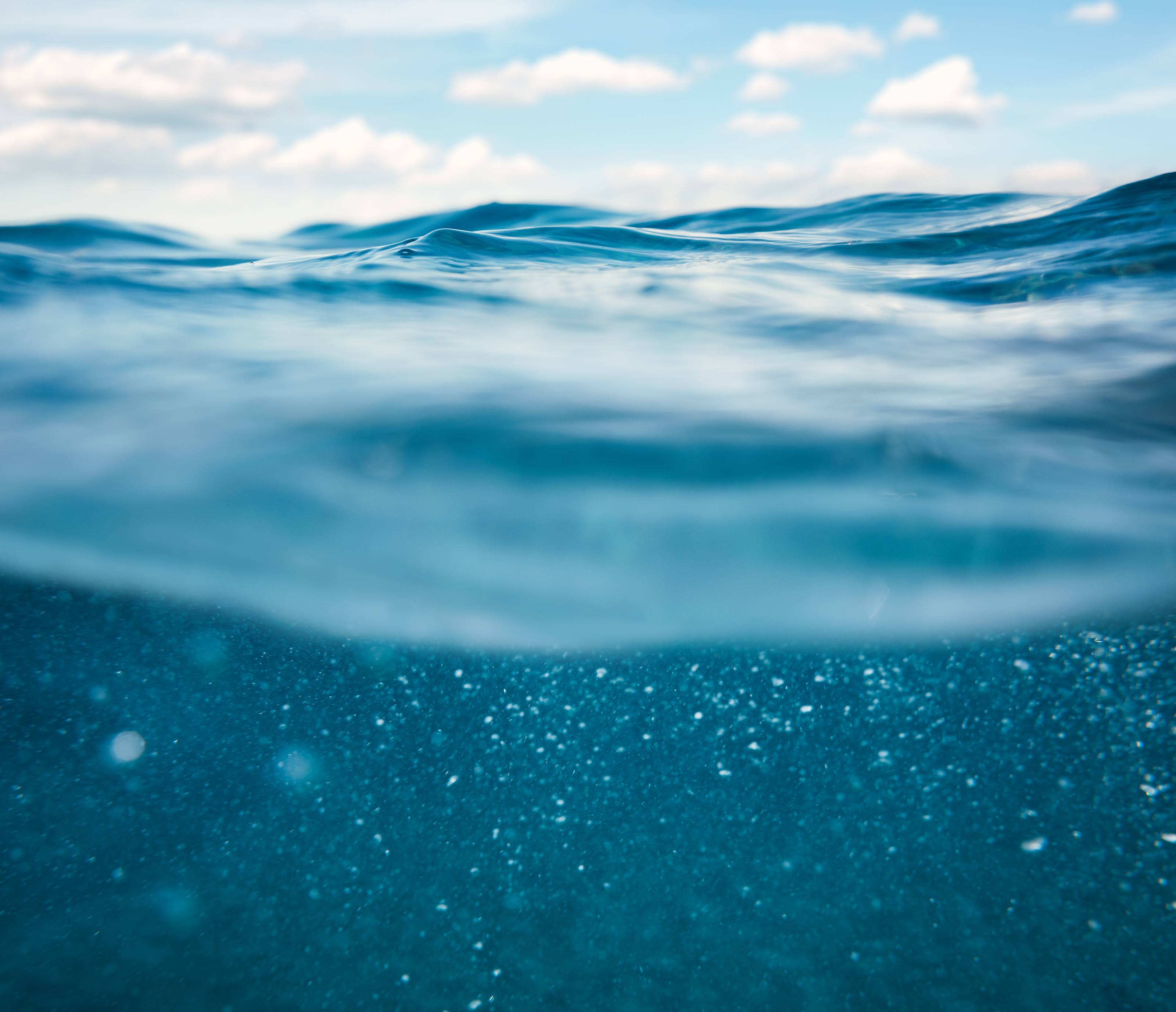 Blau-schimmerndes Wasser im Ozean Blau-schimmerndes Wasser im Ozean unter hell-blauem Himmel mit Wolken.