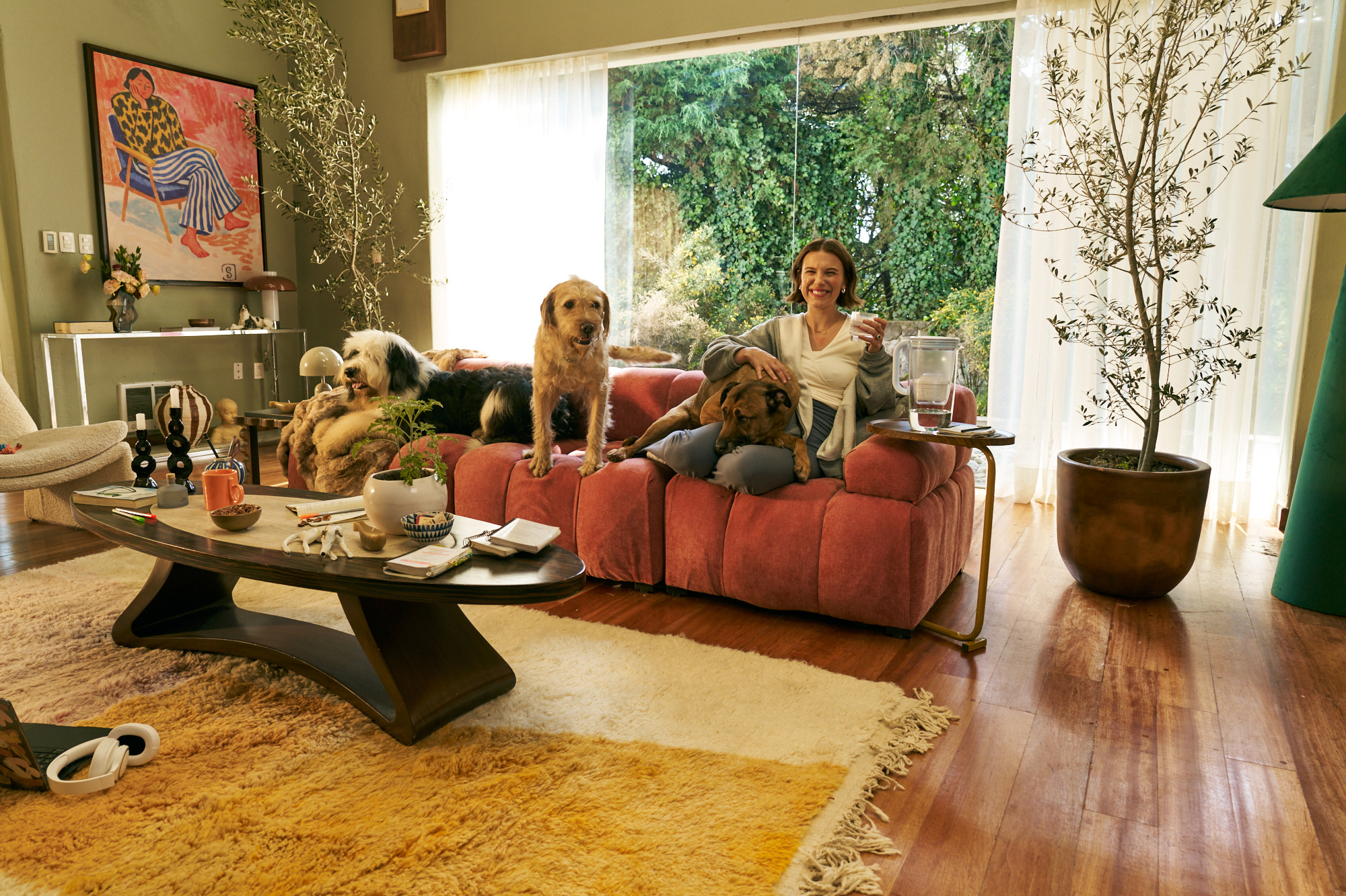 Millie Bobby Brown sitzt auf Sofa mit drei Hunden, Glastisch vorn und BRITA Karaffe am Fenster.