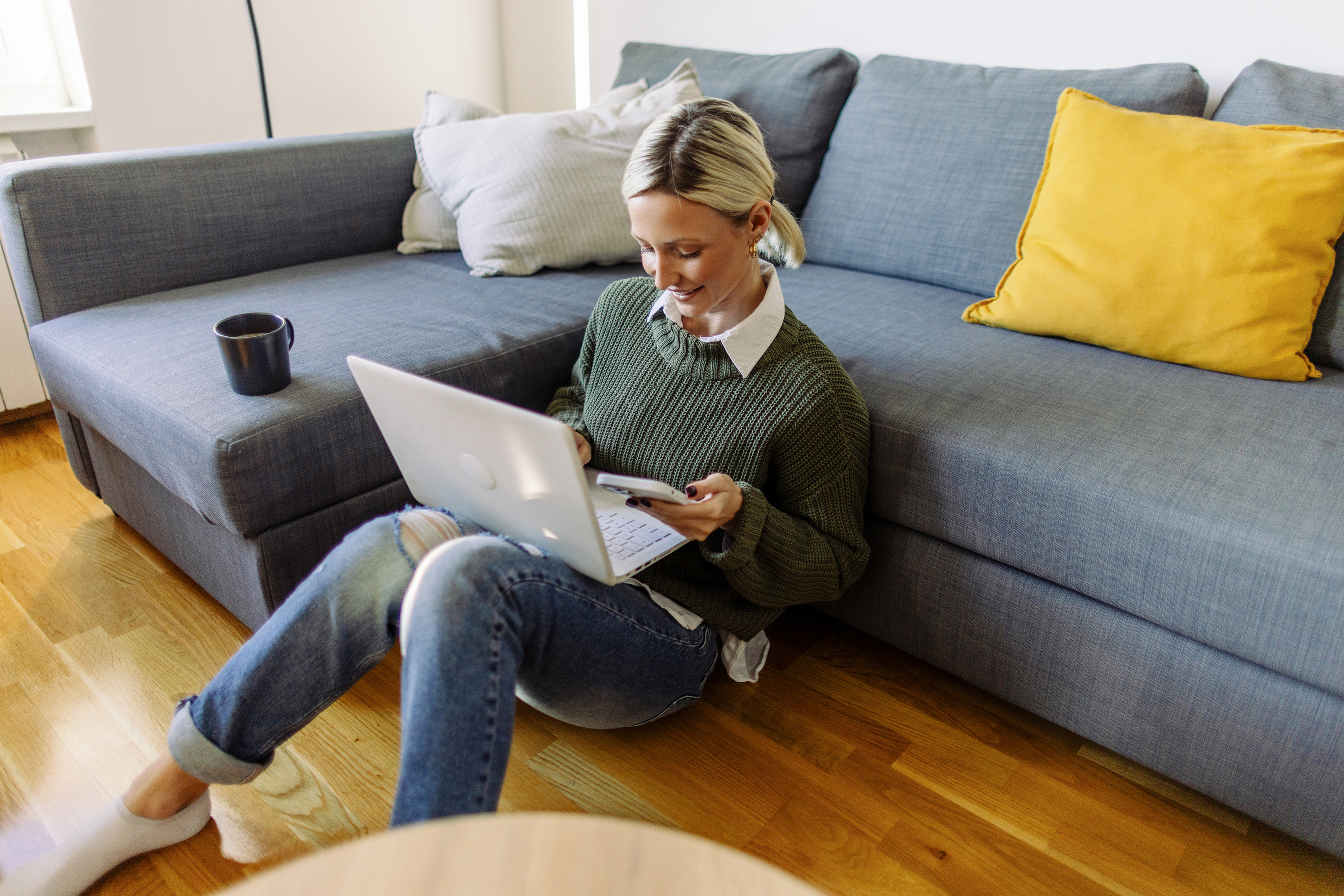 Eine junge Frau sitzt lächelnd mit ihrem Laptop und Smartphone auf dem Boden vor der Couch.