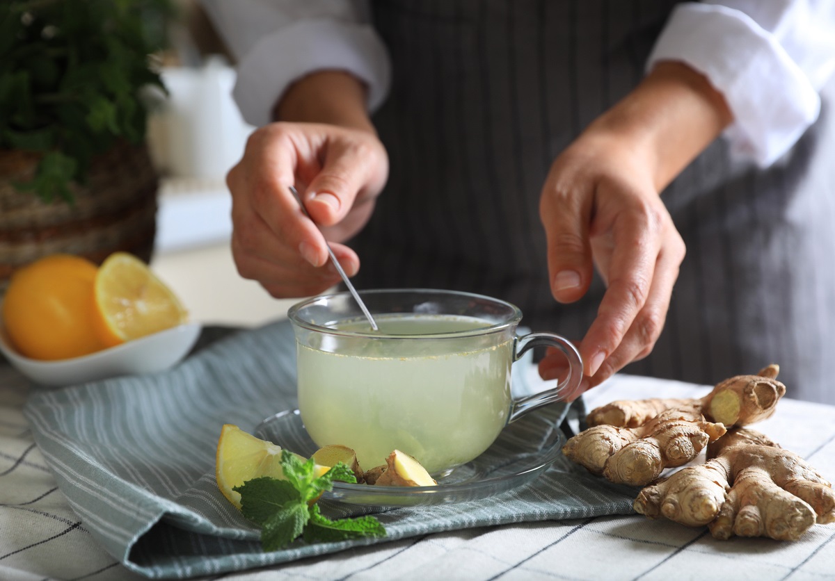 Mann rührt mit einem Löffel Detox-Getränk in einer Glastasse um, Ingwer liegt daneben auf dem Tisch