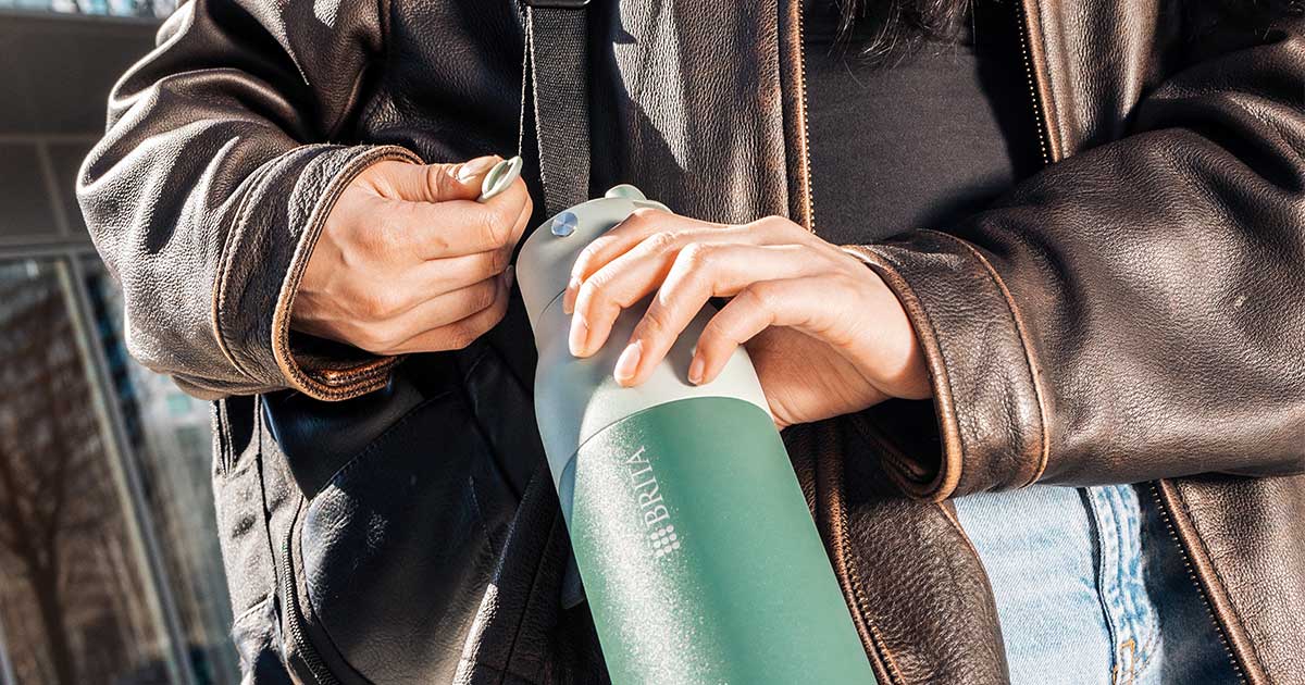 Freie Hände unterwegs – BRITA LARQ Filterflasche Frau befestigt BRITA LARQ Filter-Trinkflasche am Rucksack.