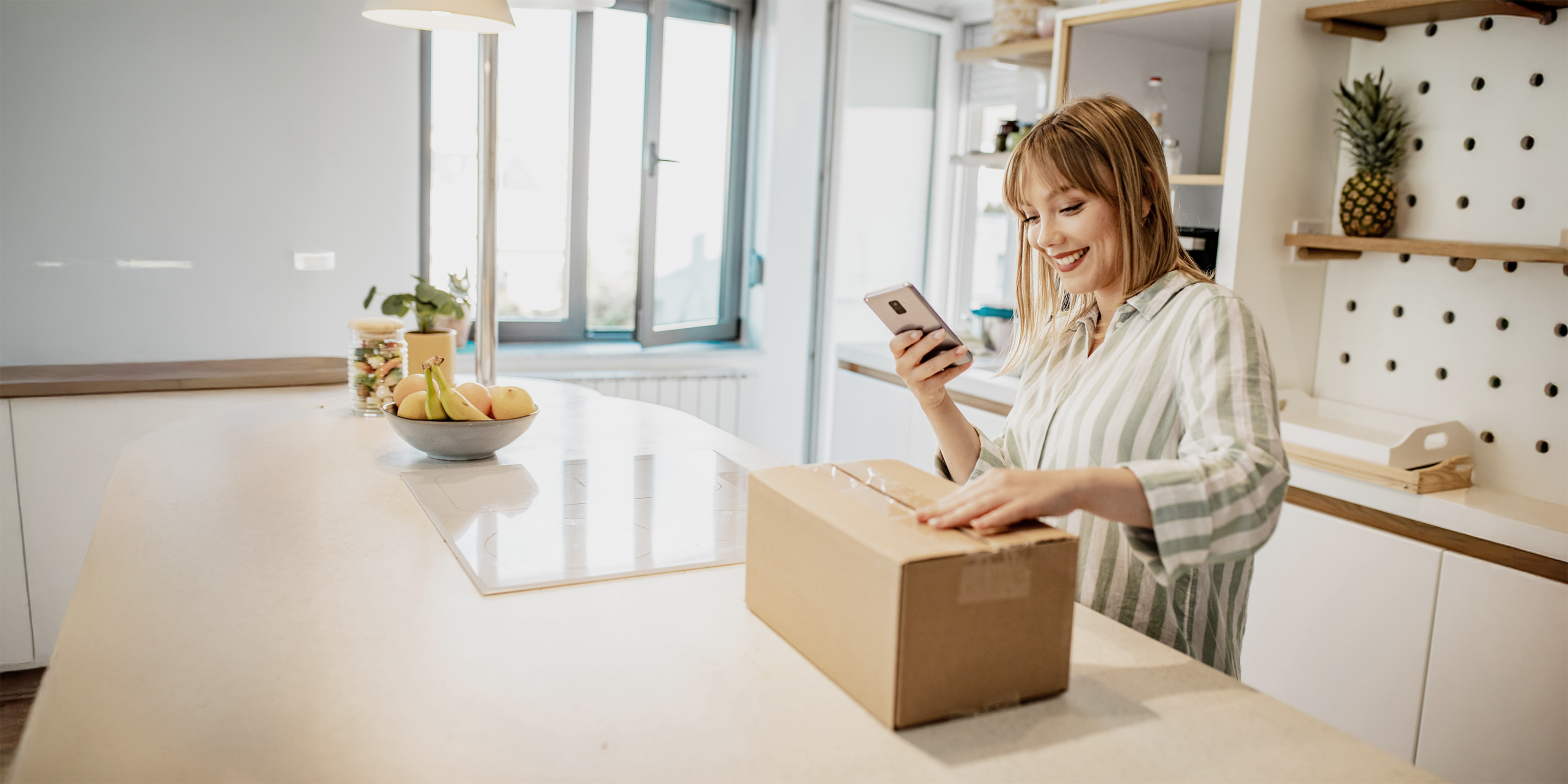 Junge Frau in Küche öffnet freudig ein Paket, in der Hand hält sie ein Handy