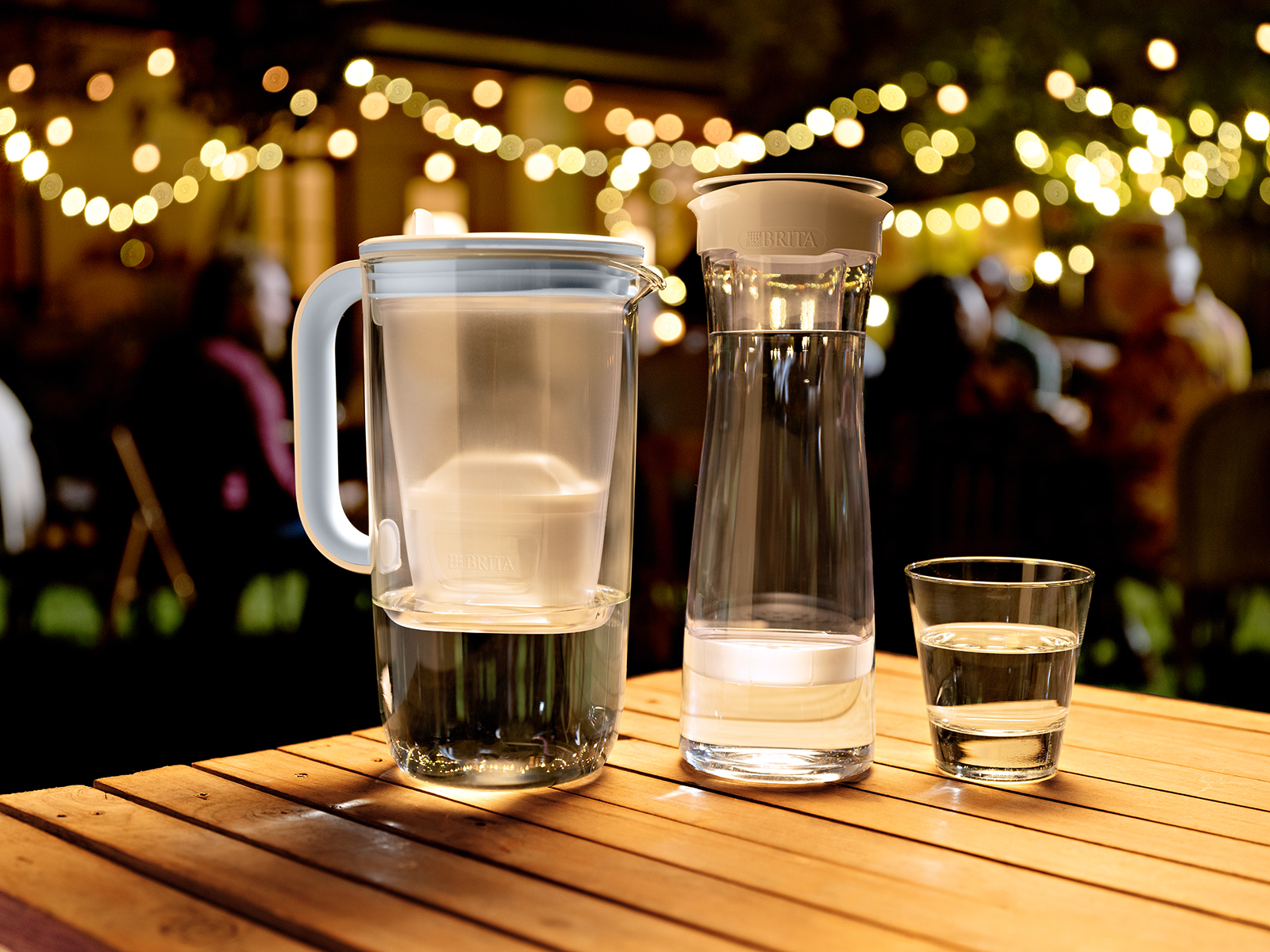 Zwei Tischwasserfilter aus Glas stehen auf einem Tisch und im Hintergrund findet eine Party statt.