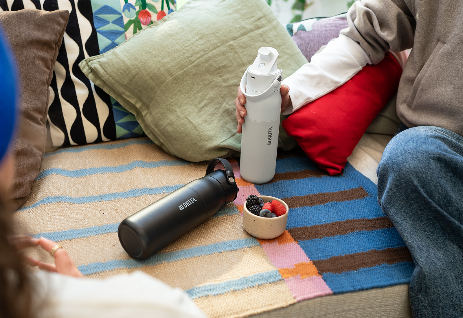 Eine weiße und eine schwarze BRITA LARQ Wasserfilterflasche auf einer Couch neben Kissen.